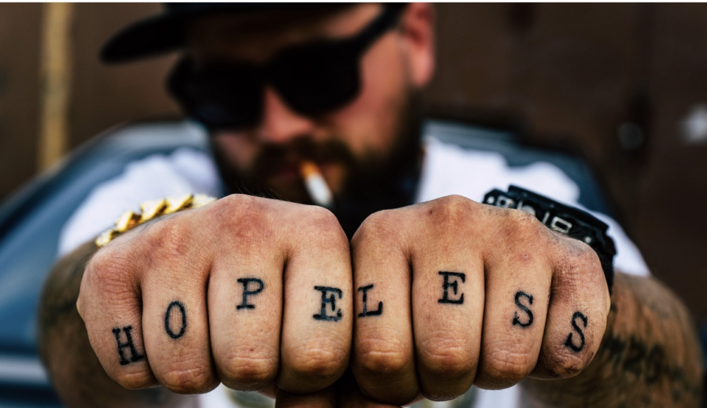 "HOPELESS" tattooed onto fingers of man smoking, wearing sunglasses and hat